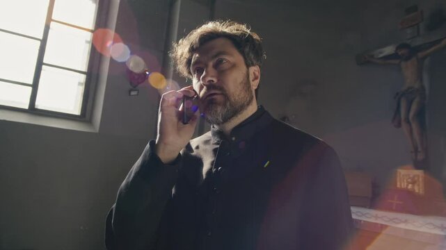 Handheld tilt up shot of middle-aged Caucasian priest in black soutane talking on mobile phone standing in church with crucifix in background, illuminated by bright light coming through window