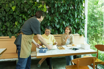 Asian senior woman and Caucasian woman sit at a cafe table, smiling as a male waiter serves tea and pastries, sharing a joyful retirement moment together., Concept small business