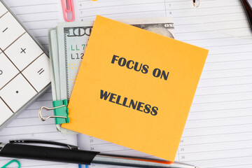 A bright yellow sticky note highlights the phrase focus on wellness. Surrounding it are money, financial tools, and stationery, showing an organized workspace connecting wellness and finance