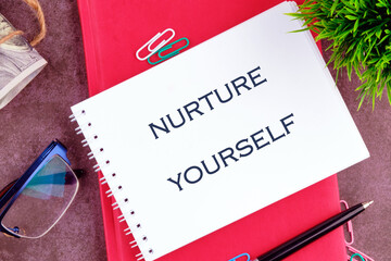 A handwritten note stating NURTURE YOURSELF is placed on a bright red notebook alongside colorful...
