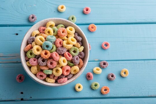 Colorful breakfast cereal loops in white bowl. Bowl of vibrant multicolored cereal loops on a bright blue wooden table, with pieces scattered around for a playful breakfast scene