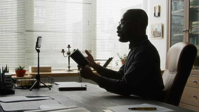 Side view of bearded bald young priest giving spiritual support to parishioner while communicating via video call holding bible book and rosary at wooden desk with phone tripod on it