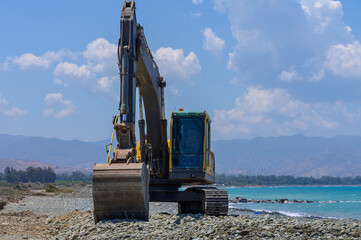 Heavy yellow excavator flattening sand on Cyprus beach. Construction machinery near sea under bright sun. Maintenance work in progress.