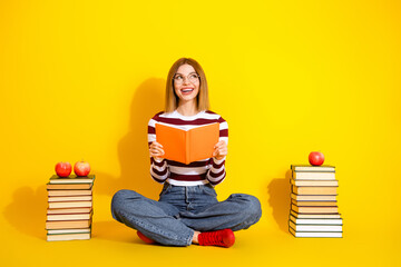 Smiling young woman sitting with book and reading on a vibrant yellow background, showcasing...