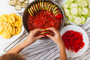 Homemade ratatouille preparation and serving. Child's hands placing vegetable slices into a baking...
