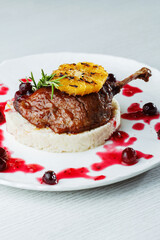 Gourmet duck leg with grilled orange, cherry sauce, and creamy risotto on a white plate, and white wooden table