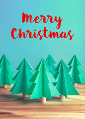 Trendy folded paper Christmas tree decorations on a wooden table against a turquoise green background that says Merry Christmas, easily replaced with your own personal message.
