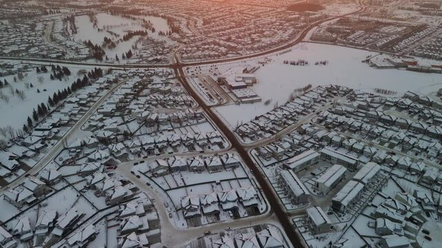 Aerial panoramic drone view of a suburban neighborhood in Edmonton, Alberta, Canada at winter sunrise. Snow-covered houses, roads, and open fields stretch across the horizon.