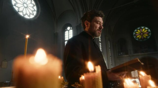 Slowmo back view shot of serious middle-aged Caucasian priest in black soutane reading Bible out loud under candlelight in Catholic church with exquisite stained glass windows behind