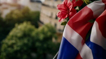 French flag waves with red flowers on a balcony overlooking blurred buildings and green trees during daytime.