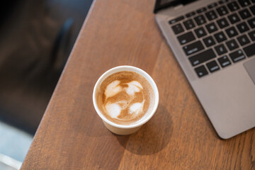 Close-up of Hot coffee latte with latte art milk foam in cup made of paper and laptop computer on wood desk office desk in coffee shop at the cafe,during business work concept