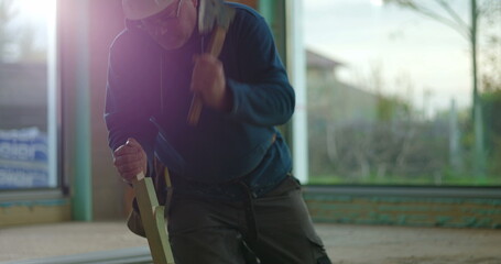 Construction worker hammering wood piece with focus and strength, kneeling on indoor construction...