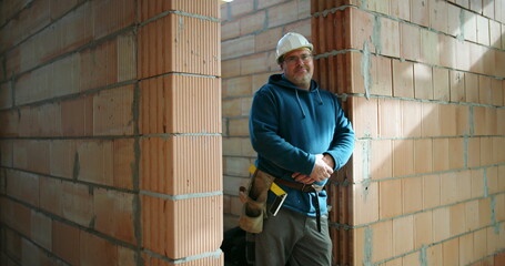 Construction worker in blue hoodie and safety helmet leaning on brick wall in building under construction, tool belt around waist, confident and relaxed posture