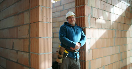 Construction worker in blue hoodie and safety helmet leaning on brick wall in building under...