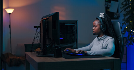 A focused young woman plays online multiplayer games with teammates in a dimly lit room. He wears a...