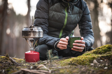 Person mit Tasse in der Hand vor Campingkocher in der Natur
