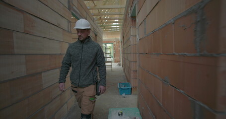 Fototapeta premium Construction worker wearing helmet walking through narrow hallway of unfinished house, passing exposed brick walls and ceiling beams in residential construction site