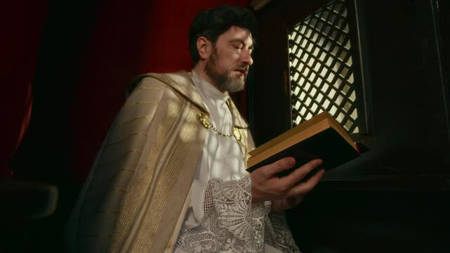 Slowmo shot of middle-aged Caucasian priest in golden cope sitting inside confessional booth and reading Bible out loud through latticed screen to parishioner in church