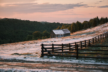 Serene sunset over a snowy field, with a rustic wooden barn and fence. Peaceful rural landscape,...