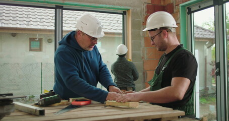 Construction workers measuring and preparing wood on workbench, teamwork and precision, safety helmets, focused on carpentry and building tasks, indoor construction site