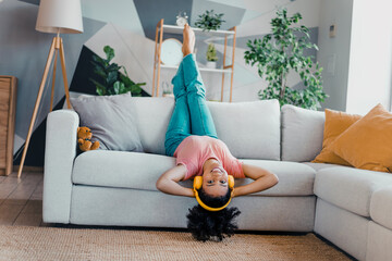 Lovely woman relaxing upside down on a couch while enjoying music with headphones in a bright cozy...