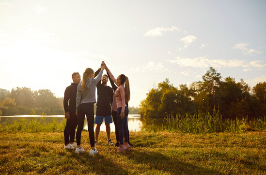 Happy sporty group of people standing in a circle holding hands up in stack on the lake shore in the morning after sport training in the park. Workout in nature and healthy lifestyle concept.