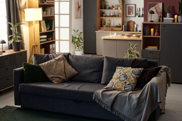 Cozy living room featuring a comfortable gray sofa adorned with patterned pillows and throw. Modern kitchen area visible in the background with some fresh fruits placed on counter