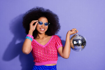 Young woman with afro hairstyle wearing trendy clothing holding a disco ball on a violet background...