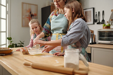 Mother and two daughters baking in kitchen, measuring and mixing ingredients, showing family bonding and teamwork, enjoyed moment of togetherness, kitchenware visible