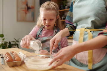 Young blonde girl in pink apron carefully measuring flour while baking with an adult in home...