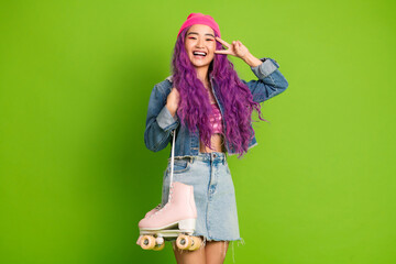 Cheerful young woman with purple hair and pink roller skates posing with peace hand gestures against a green backdrop