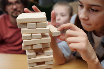 Child meticulously removing block from Jenga tower while adults watch attentively in background,...