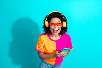 Cheerful young woman with headphones and vibrant outfit holding smartphone on a turquoise background
