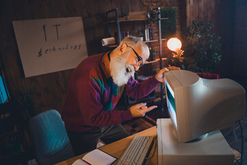 Elderly man working with vintage computer in a cozy office at night, symbolizing technology...