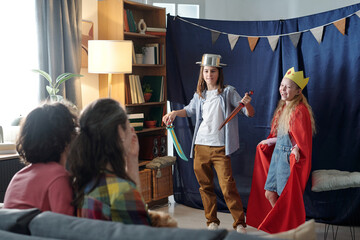 Children dressed as characters performing play on living room stage. Family members sitting and enjoying performance with books and decorations in background