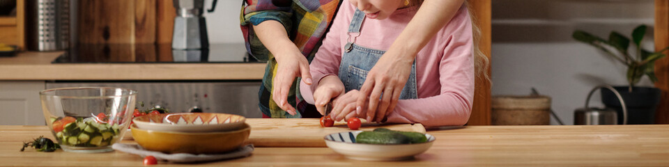 Child and adult preparing a healthy vegetable meal on wooden kitchen counter with various kitchen tools and fresh ingredients visible. Emphasis on involvement and learning
