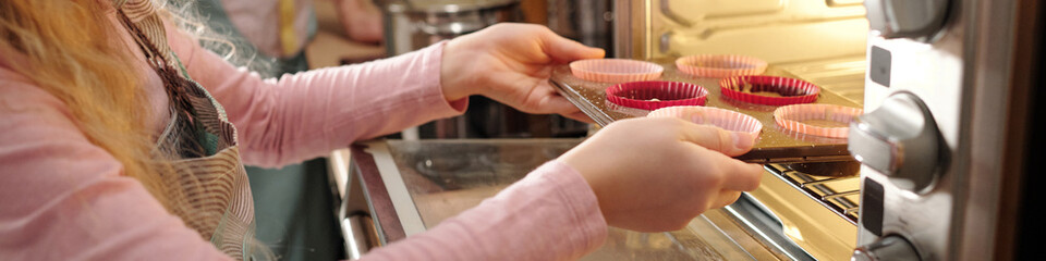 Child with long hair baking cupcakes in oven, using pink silicone molds and wearing apron, focused on placing the tray in oven