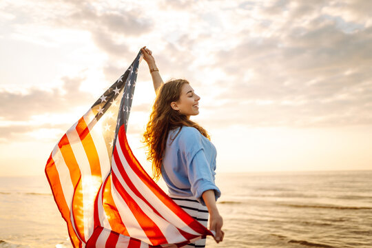 A joyful woman has fun walking along the beach with an American flag at sunset. A young woman is happy and enjoying nature with a US flag. Freedom and enjoyment concept.
