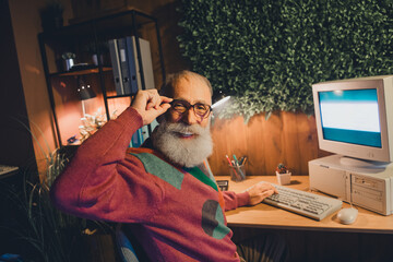 Elderly man with beard working on classic computer wearing sweater in stylish home office, smiling...