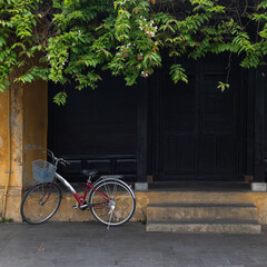 An ordinary red bicycle with a basket in front stands in the shade of foliage. In the background is the shabby facade of a yellow house and dark wooden doors and shutters.