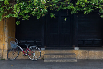 An ordinary red bicycle with a basket in front stands in the shade of foliage. In the background is the shabby facade of a yellow house and dark wooden doors and shutters.