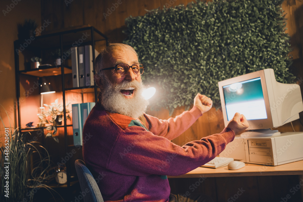 Poster elderly man joyfully working with a computer indoors in a cozy evening setting, showcasing freelance