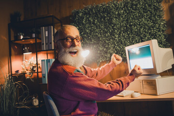Elderly man joyfully working with a computer indoors in a cozy evening setting, showcasing...