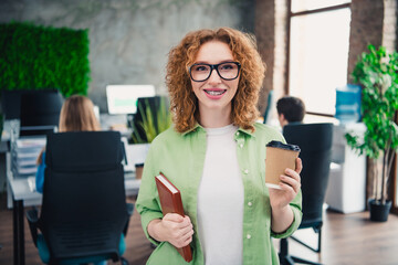Smiling redheaded woman in a casual office setting holding a coffee and notebook, radiating...