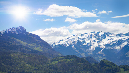 Panoramic alpine landscape with snowy mountain peaks and lush green valleys in springtime. Bright clouds and blue sky complete the peaceful nature scene