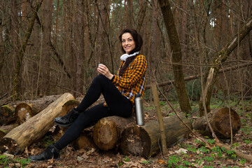 Beautiful happy emotion woman hiking in the forest relax sitting on logs looking up on sunny spring weather. Girl rest enjoy, drinking from cup of thermos in style fashion clothes in woods..