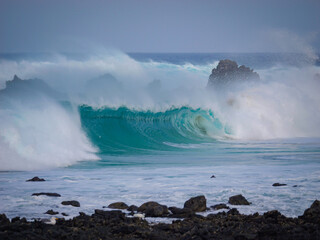 Big closeout wave, lifted by an offshore wind, collapses onto a volcanic shore. Misty spray rises above the turquoise wave into the air in beautiful arc. Raw energy and beauty of Atlantic Ocean.
