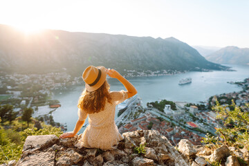 A young woman on a rock enjoys the view of the bay. Tourist in a hat feels free and has fun on the...