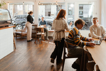 Two White female cafe workers are attending to a multinational group of people inside a cafe with large windows, wooden furniture, a pastry display counter, potted plants, and a cozy urban atmosphere.