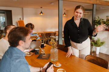 A young White female cafe owner in a black shirt and beige trousers smiling and greeting a group of male customers seated at a wooden table in a cozy cafe interior with drinks and pastries.
